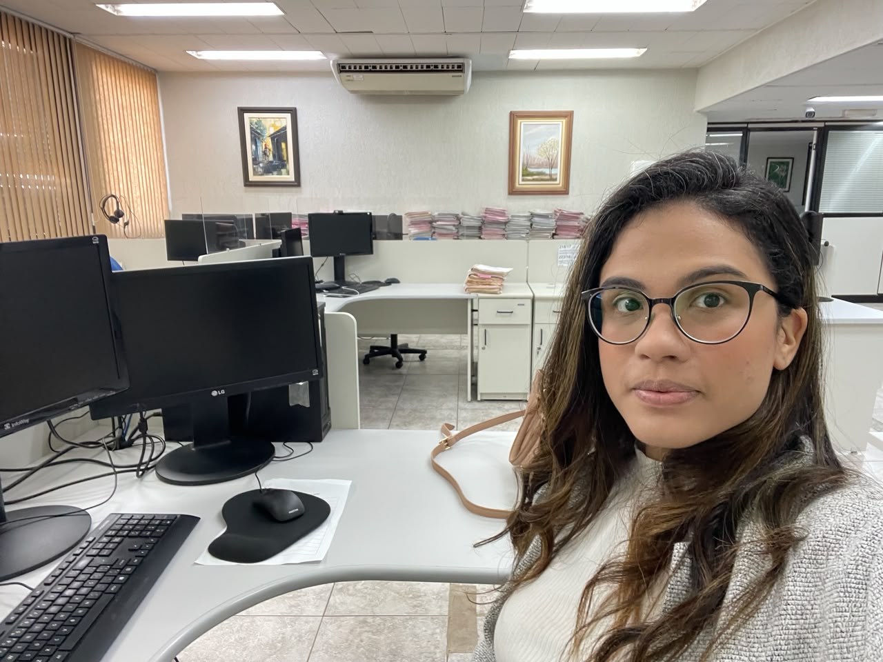 Pâmela na mesa de trabalho da Justiça Federal, com monitores à frente e pilhas de processos organizados ao fundo